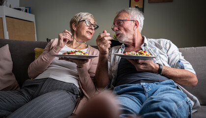 Senior couple sitting on sofa and eating pasta. They're having pleasant conversation and joying in...