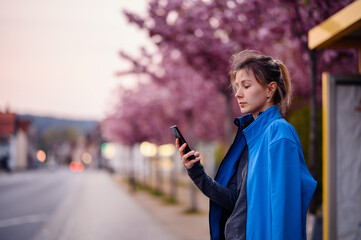 Young girl in blue jacket and jeans standing on bus stop with sakura alley looking on watch on hand