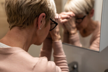 Senior woman standing in front of the mirror and looking at her reflection. Depression concept.