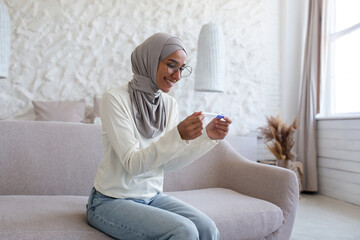 Photo from the side. Smiling young Muslim woman in hijab sitting on sofa at home and holding disposable and quick pregnancy test. Happy with the positive result.