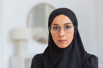 Close-up photo. Portrait of a young woman, an immigrant, a refugee from eastern countries, wearing a black hijab and glasses, looking seriously into the camera.
