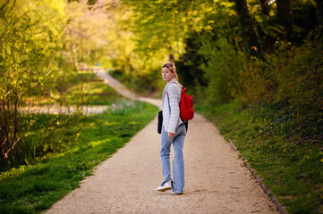 Young blonde girl in spring outfit walks in green park with red backpack and laptop