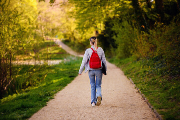 Obraz premium Young blonde girl in spring outfit walks in green park with red backpack and laptop