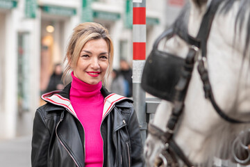 Street portrait of a blonde woman, looking at a horse-drawn carriage in a European city. Concept: tourism and travel, horseback riding.