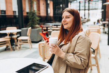 Young woman enjoying a coffee, sitting on the cafe terrace on the modern city street. Person sitting at table and using smartphone outdoors. Online education, order, working or shopping concept