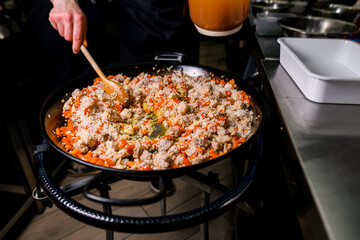 Chef stirs rice, meat and vegetables using wooden spatula in frying pan