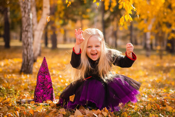 A little girl with blond hair in a black dress and a hat in the image of a witch for Halloween in...
