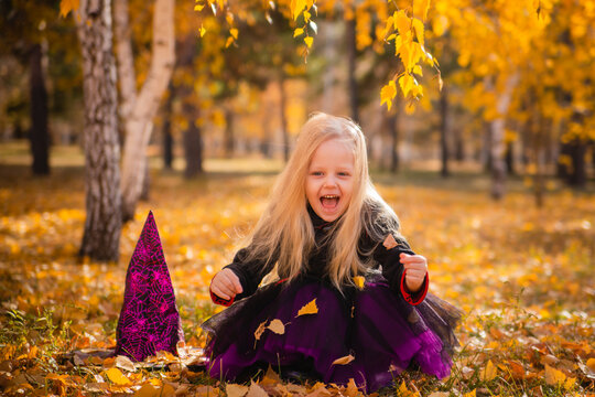 A little girl with blond hair in a black dress and a hat in the image of a witch for Halloween in autumn in a park among the leaves.