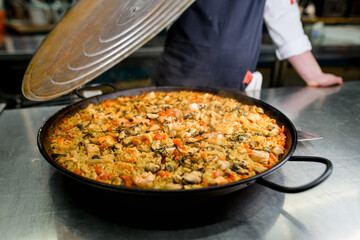 Chef holding pan lid in hand and presenting traditional Paella with shrimp, mussels, vegetables.