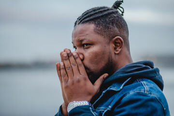 A black young man with dreadlocks in a blue denim jacket and sweatpants is standing on the embankment.