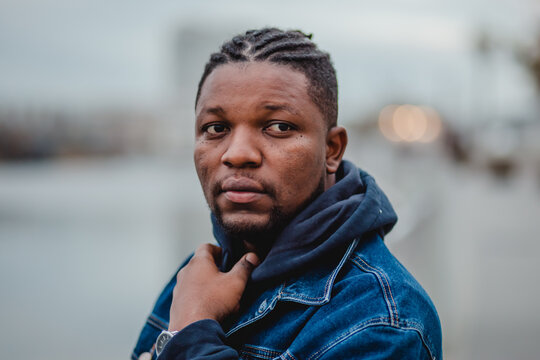 A black young man with dreadlocks in a blue denim jacket and sweatpants is standing on the embankment. - Powered by Adobe