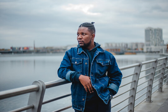 A black young man with dreadlocks in a blue denim jacket and sweatpants is standing on the embankment.