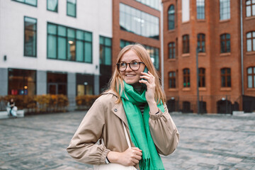 Fototapeta premium Beautiful young woman talking on smartphone walking through city street. Portrait of gorgeous smiling female using mobile phone. High quality photo