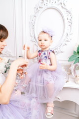 Baby girl elegant dress. A one-year-old girl in a puffy dress and a cute bow poses against the backdrop of a bright room with a dressing table and flowers.