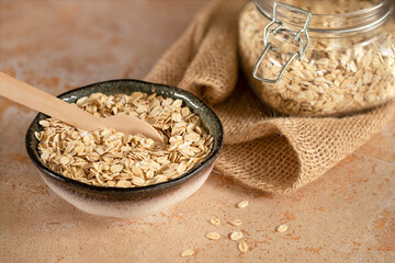 Oat flakes, cereals in a handmade ceramic bowl on burlap on a beige stone table, a spoon and a glass jar with oatmeal