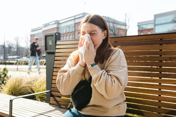 Unhappy caucasian millennial red-haired woman sneezing in napkin and siting in park bench, Young...