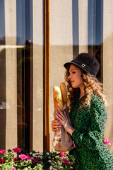 Pensive woman in French style near window with baguettes in hands, thought looking away. Pretty woman in green dress and hat enjoy resting outdoors. Summertime relaxing concept. Copy ad text space