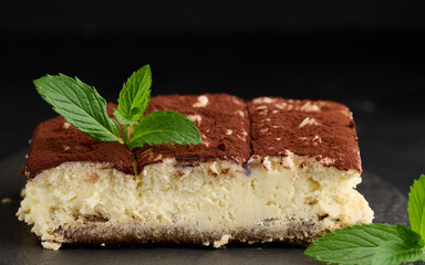 A piece of tiramisu sprinkled with cocoa, on top of a sprig of fresh mint on a black background