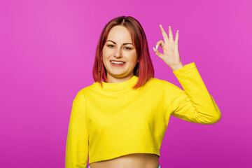 Portrait of happy and positive woman smiling carefree and shows ok sign, standing in yellow t-shirt on pink background