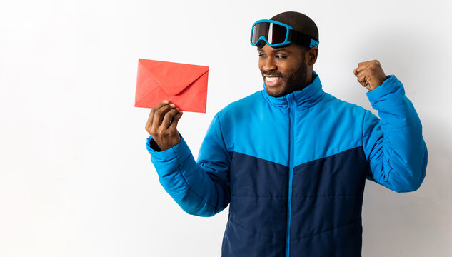 Snowboarding dark-skinned man wears blue suit and ski goggles. The male shows a gift envelope for a store isolated on a white background while looking at it from the side. Winter concept.