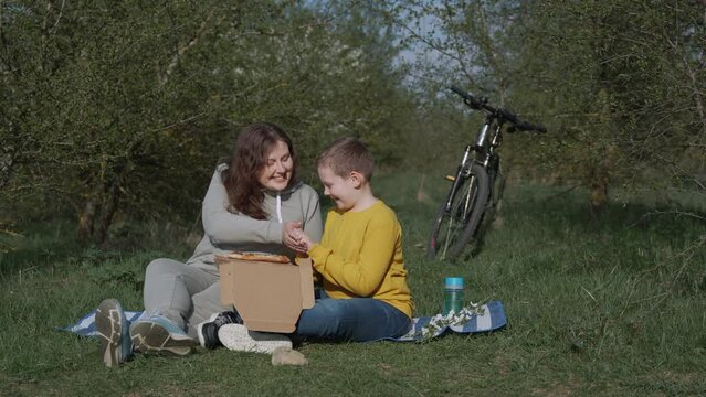 Mother and son are relaxing on lawn near trees with bicycle on spring day. Child tries to take pizza, woman playfully pushes away hands. Happy family smiling, resting at picnic after bike ride.
