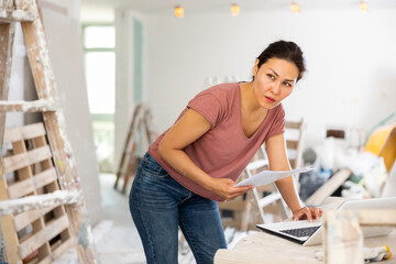 Asian woman construction manager checking project documentation and using laptop while working in apartment.