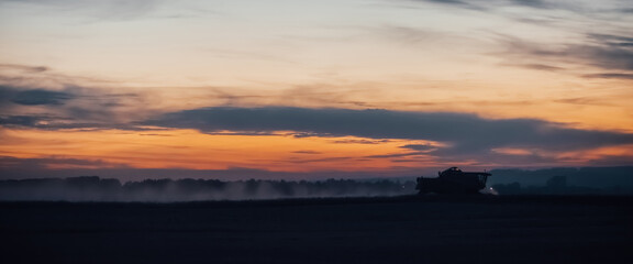 Naklejka premium Silhouette of harvester machine to harvest wheat on sunset. Combine harvester driving on field on sunrise. Beautiful dawn sky above wheat field. Combine working in dusk. Wonderful twilight landscape.