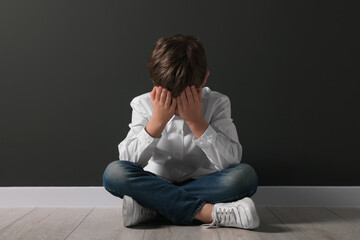 Upset boy sitting on floor near black wall. School bullying