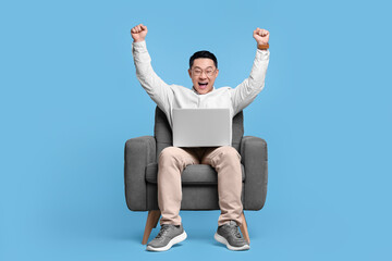 Emotional man with laptop sitting in armchair on light blue background