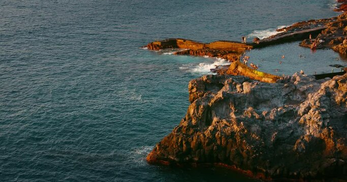 People swim in natural pool in the Atlantic ocean of Tenerife at sunset. Charco de Isla Cangrejo. Historical landmark. Piscina natural.