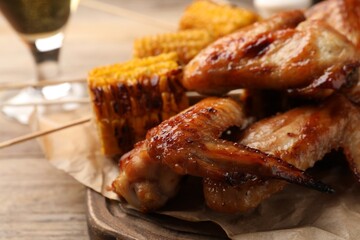 Delicious baked chicken wings and grilled corn served on wooden table, closeup