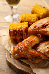 Delicious baked chicken wings and grilled corn served on wooden table, closeup