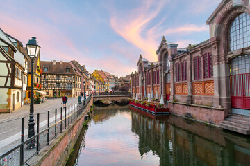 Fototapeta premium Picturesque half timber buildings across from the riverfront Marché Couvert covered market at sunset along the Lauch River in the Petite Venice area of Colmar, France.