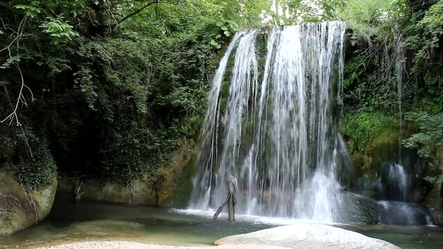 Basking view at Cascata "de lu Vagnat&ograve;" in Sarnano with the waterfall's silvery waters flowing down in threads, washing thick vegetation and robust rocks, splashing and collecting in an emerald basin