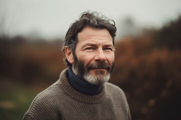 Fototapeta premium Portrait of a handsome middle-aged man with gray beard and mustache, wearing a warm knitted sweater, standing in the autumn park.