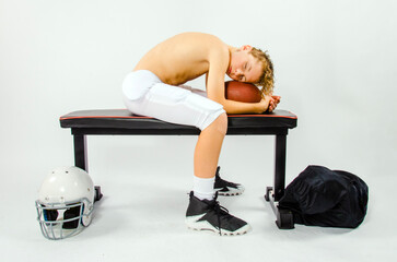 Youth male football player asleep shirtless on bench with his head resting on a football