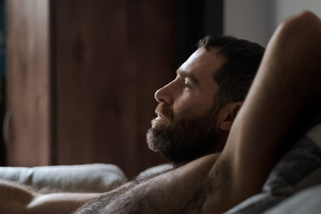 Leaning back shirtless on the sofa, a handsome man expresses being totally relaxed during a casual day at home. His thick beard, chest hair, and underarm hair are part of his look.