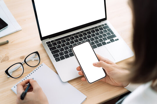 Lady uses smartphone with blank screen and writes in blank notepad in front of laptop with white screen on wooden table.