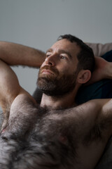 Leaning back shirtless on the sofa, a handsome man expresses being lost in thought, daydreaming while relaxing at home His thick beard, chest hair, and underarm hair are part of his look.