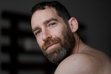 Looking over his shoulder while relaxing at home, a man expresses a coy smile and masculine calm. He is shirtless, with a thick beard that draws attention to his face.