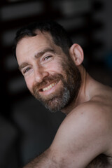 Looking over his shoulder while relaxing at home, a man expresses a joyful smile and direct eye contact. He is shirtless, with a thick beard that draws attention to his face.