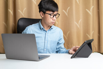 Happy young Asian businessman or investor using laptop, tablet for trading, investing on new cryptocurrency as Bitcoin or stock trading platform at home. Young money investing and trading concept.