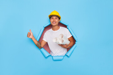 Smiling man in yellow hard hat and white T-shirt posing inside the cutout of blue background, holding rolls of paper in his hand, thumbs up with other hand, good offer concept, copy space, high