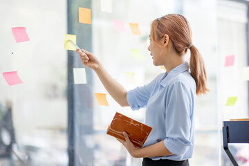 Young business asian woman creative team using post it notes in glass wall to writing finance...