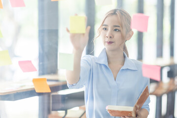 Young business asian woman creative team using post it notes in glass wall to writing finance expert strategy business plan to development grow to success.