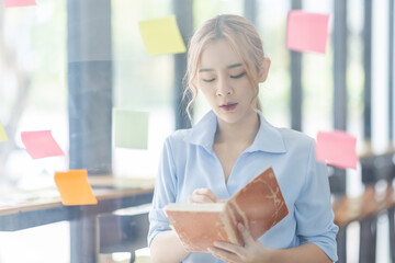 Young business asian woman creative team using post it notes in glass wall to writing finance...