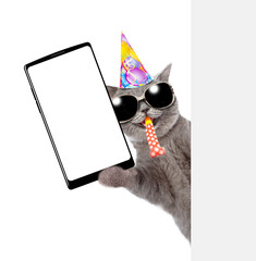 Happy cat wearing sunglasses and party cap blows in party horn and look from behind empty white banner  and shows big smartphone with blank screen. isolated on white background