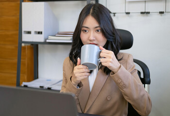 Young female worker using laptop in office, Officer holding coffee/tea mug while looking at open laptop computer on table.Cozy office workplace, online work.Businesswoman drinking coffee