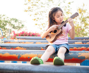 Little asian child girl play the ukulele, in the garden on the Steel pipe
