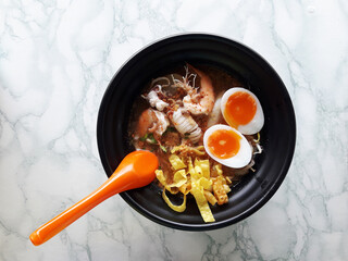 Noodles in sour soup with prawn with squid and boiled egg cut in half, Noodle in black bowl with orange spoon on white and gray background